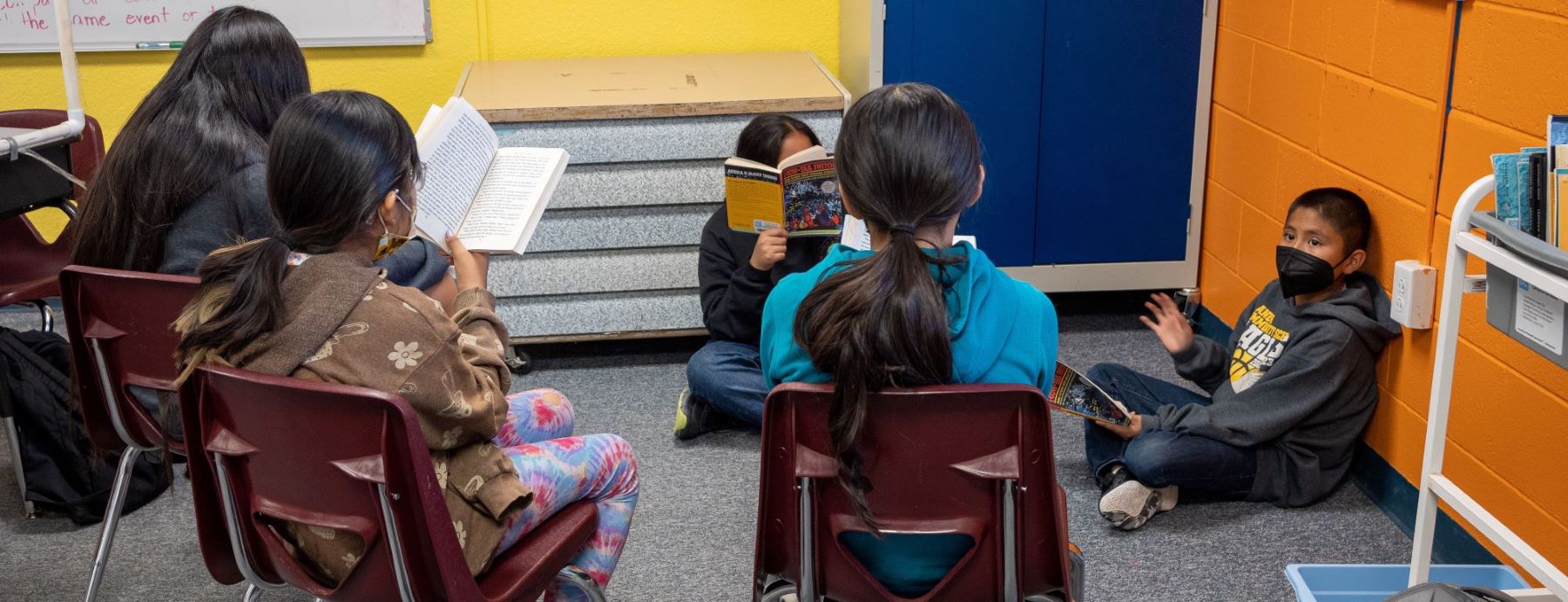 A group of students sits in a circle. Some students sit on the floor, some in chairs. Each focused on their own book in a cozy learning environment.
