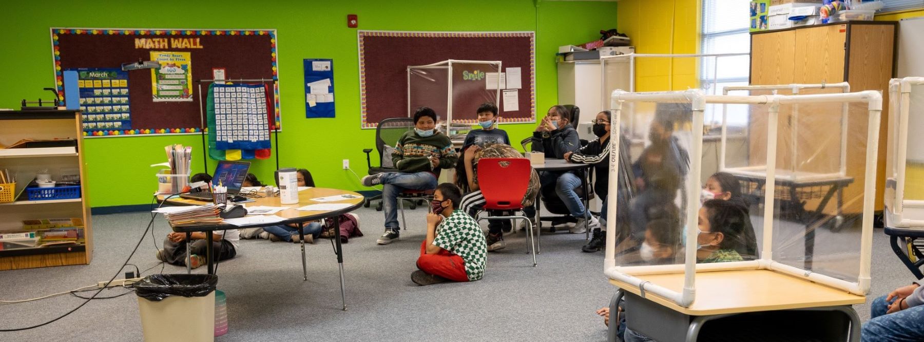 A classroom filled with students sitting in chairs and on tables, engaged in various activities and discussions.